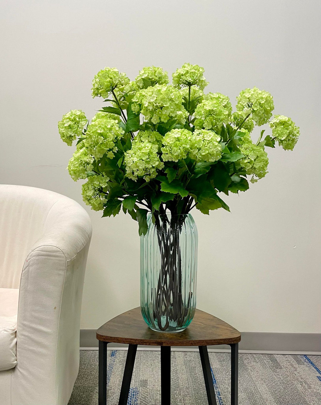 Artificial green hydrangea flowers arranged in a tall, fluted glass vase on a small wooden side table next to a cream-colored armchair. What are some stylish ways to display artificial flowers in the living room?
