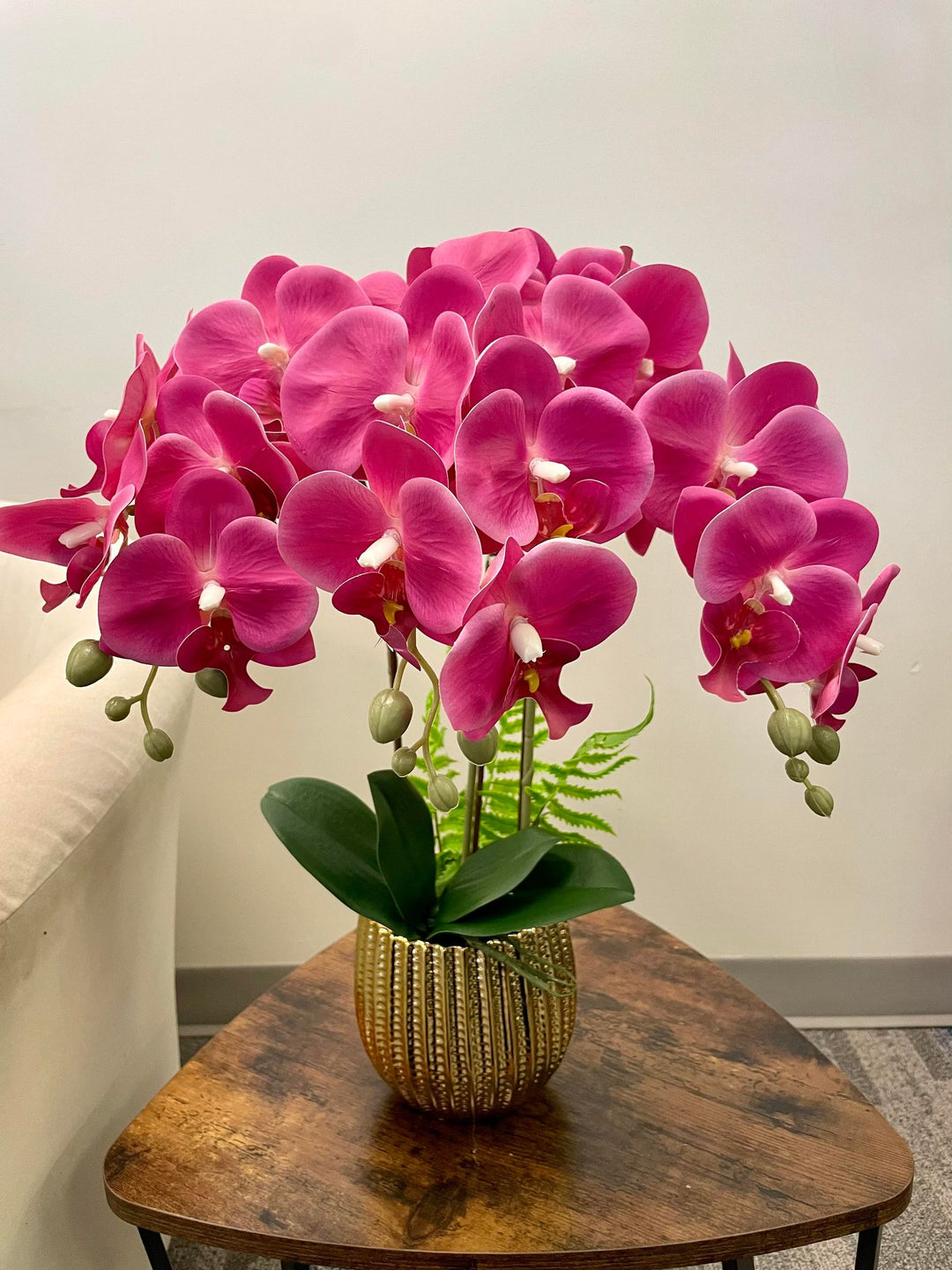 A vibrant arrangement of artificial pink orchid flowers with lush green leaves, displayed in a textured gold planter on a wooden coffee table. Frequently asked question: How can I choose artificial flowers that look lifelike and add elegance to my space?