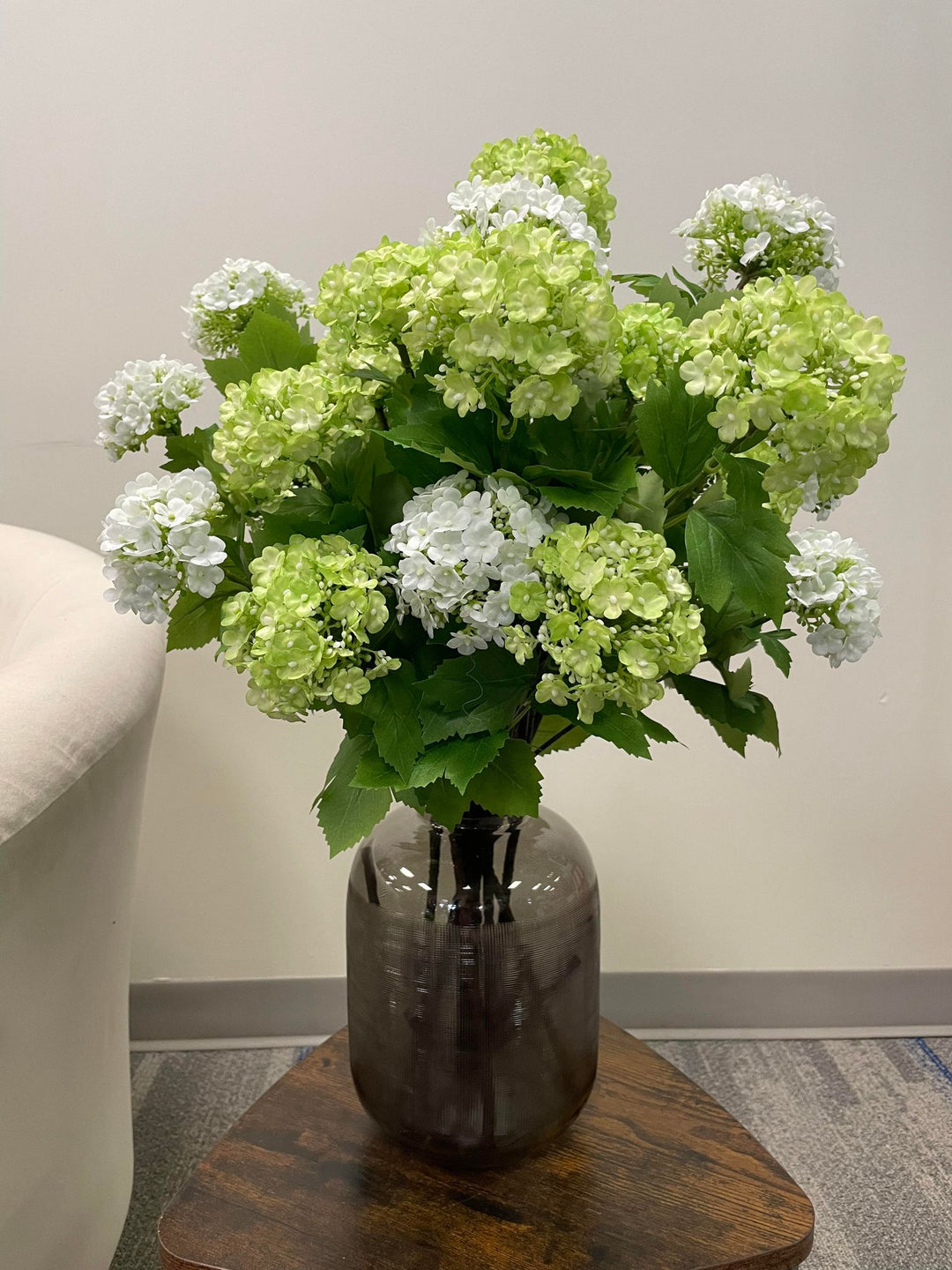 Artificial green hydrangea flowers arranged in a tall, fluted glass vase on a small wooden side table next to a cream-colored armchair. What are some stylish ways to display artificial flowers in the living room?