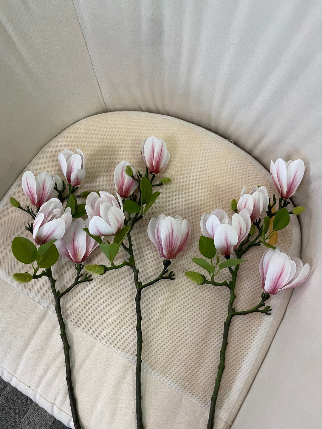 Four artificial white magnolia flower stems placed on a soft cream cushion. The stems are adorned with delicate petals and green leaves, offering a lifelike representation of magnolias, ideal for home floral arrangements or decor.