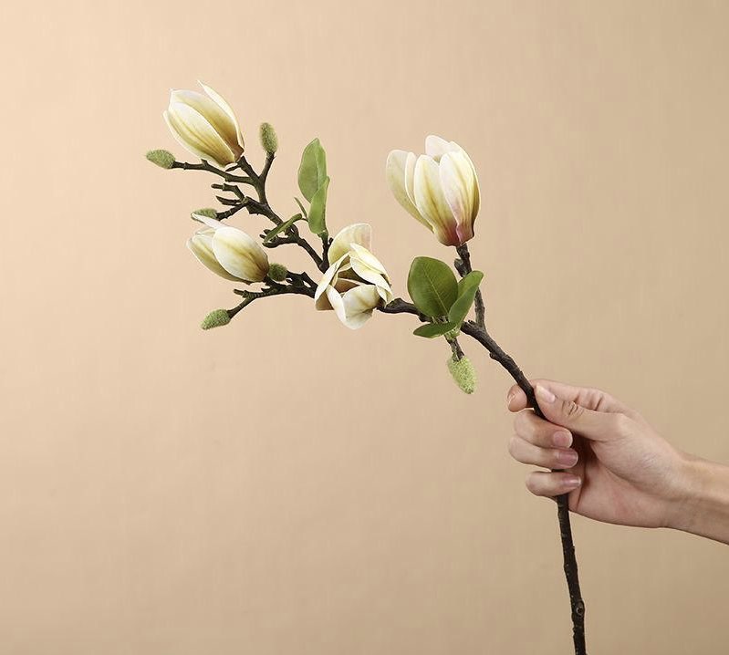 A hand holding an artificial magnolia stem with yellow and cream blossoms, showcasing realistic details. Frequently asked question: What are the best artificial flowers for adding elegance to a minimalist home design?