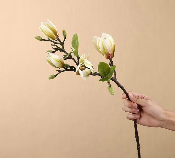 A hand holding an artificial magnolia stem with yellow and cream blossoms, showcasing realistic details. Frequently asked question: What are the best artificial flowers for adding elegance to a minimalist home design?