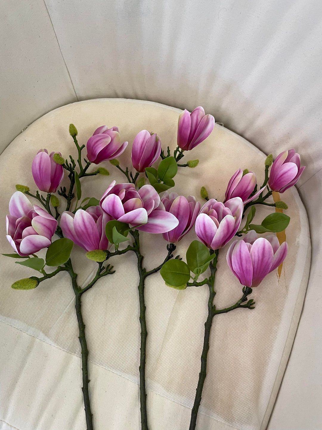 A set of artificial pink magnolia flower stems laid out on a cushioned surface, showcasing their delicate blooms and green leaves. The long stems add a natural feel to the soft, elegant arrangement.