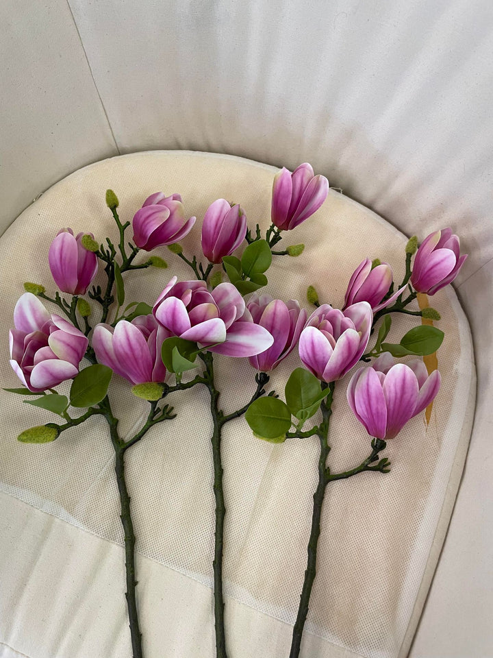 Four artificial magnolia stems with pink and white petals arranged on a light-colored chair cushion. Frequently asked question: How can I arrange artificial magnolia stems for a natural and realistic floral display?