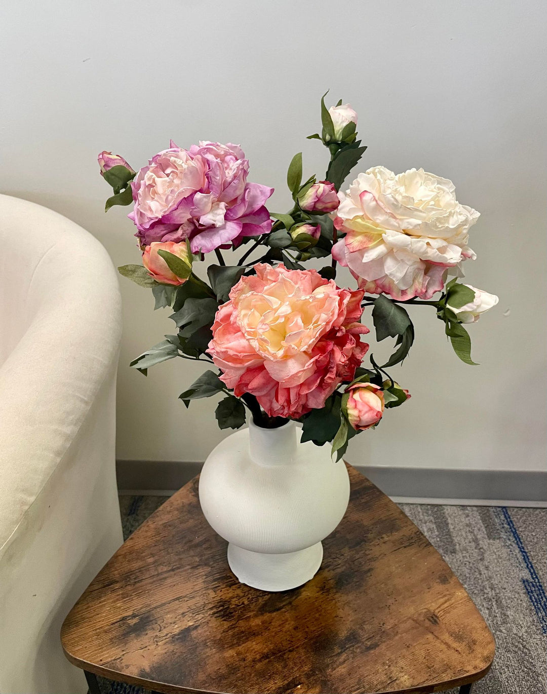 A beautiful arrangement of artificial roses in soft shades of purple, peach, and cream displayed in a white vase. The flowers are placed on a rustic wooden table next to a white armchair, creating a cozy, inviting corner.