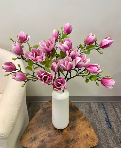 A large bouquet of artificial pink magnolia flowers arranged in a sleek white vase on a wooden table. Common question: How do artificial magnolia flowers compare to real ones for indoor home décor in terms of maintenance and visual appeal?