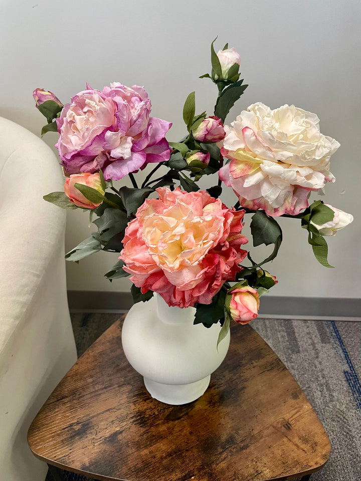 A beautiful arrangement of artificial roses in shades of peach, white, and lavender, displayed in a white ceramic vase on a wooden table. The realistic petals and buds bring a soft, romantic touch to the décor.