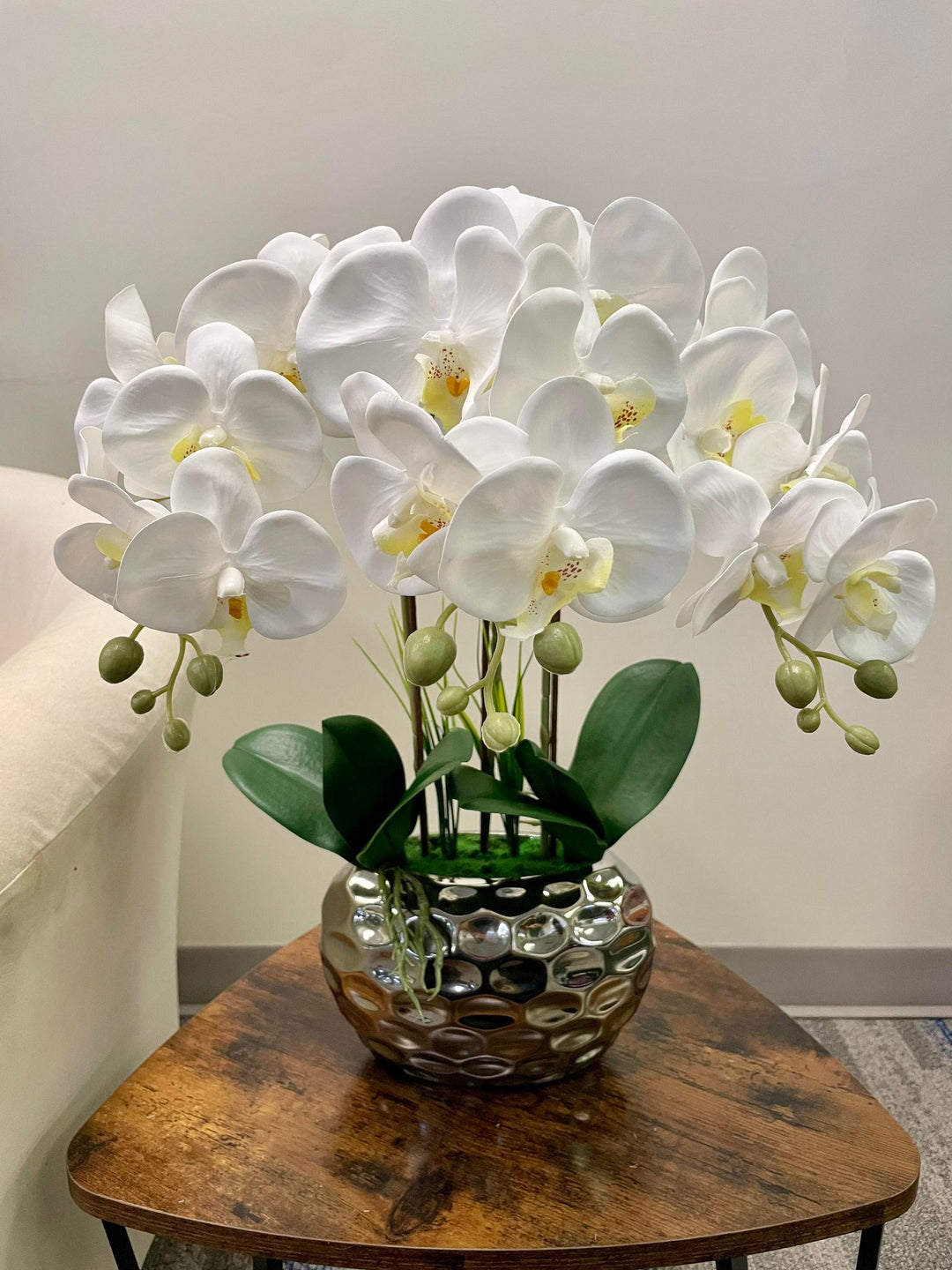 An elegant arrangement of artificial white orchids displayed in a reflective silver vase with a dimpled surface. The orchids have large, delicate petals with yellow centers and sit atop sturdy green stems with smooth, dark green leaves at the base. The vase is placed on a wooden table, with the soft, neutral background of a room, adding to the calming ambiance.