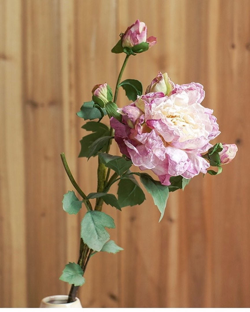 A delicate single stem of artificial purple and cream rose with buds, featuring realistic green leaves. The flower is placed against a natural wood background, offering a classic and rustic floral arrangement.