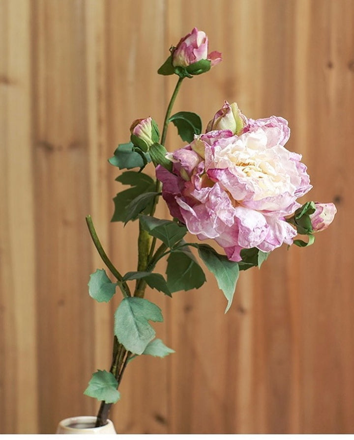 A delicate single stem of artificial purple and cream rose with buds, featuring realistic green leaves. The flower is placed against a natural wood background, offering a classic and rustic floral arrangement.