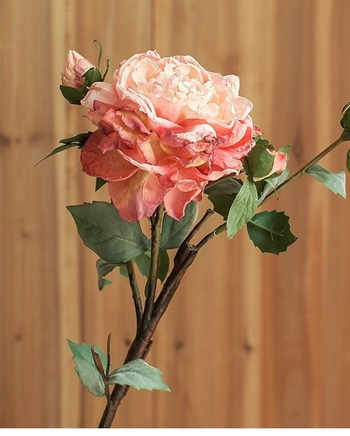 A close-up of a beautifully detailed artificial rose in soft pink and peach hues, with green leaves and a natural-looking stem. Perfect for home décor, floral arrangements, or adding elegance to any space.