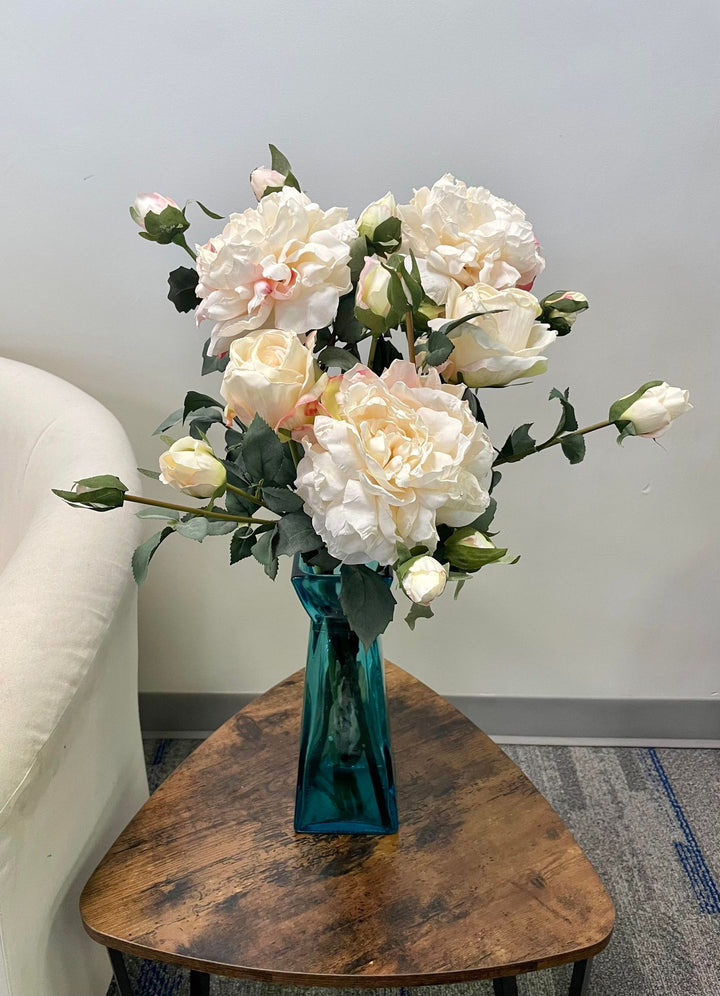 A bouquet of artificial white peonies arranged in a teal vase, sitting on a small wooden coffee table next to a cream-colored chair. Frequently asked question: How do I style artificial peonies in vases for a modern home décor setting?