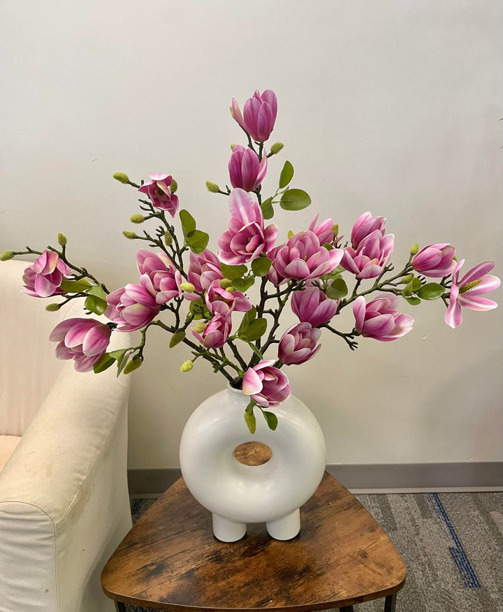 A striking display of artificial pink magnolia stems arranged in a modern white circular vase, placed on a rustic wooden coffee table next to a cream armchair. Common question: What are the best ways to style artificial flowers in contemporary home décor?