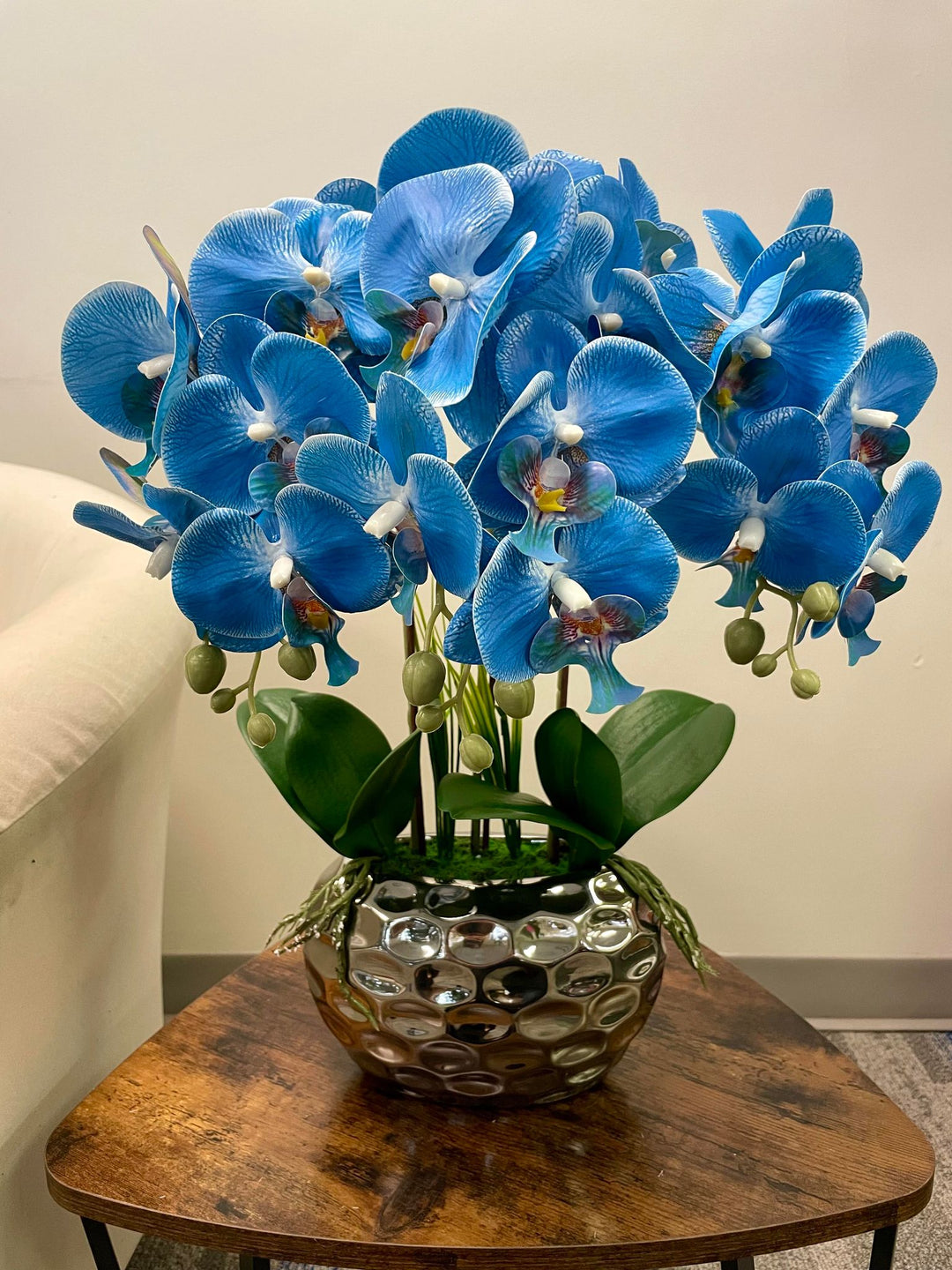 A striking arrangement of vibrant blue artificial orchids displayed in a metallic, textured vase. The rich colors of the orchids contrast beautifully with the green leaves and glossy pot, creating an elegant and eye-catching floral centerpiece.