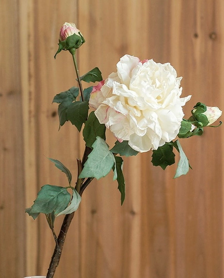 Artificial white peony stem with green leaves against a wooden background. Common question: Are artificial peonies realistic enough to replace fresh flowers in home décor?