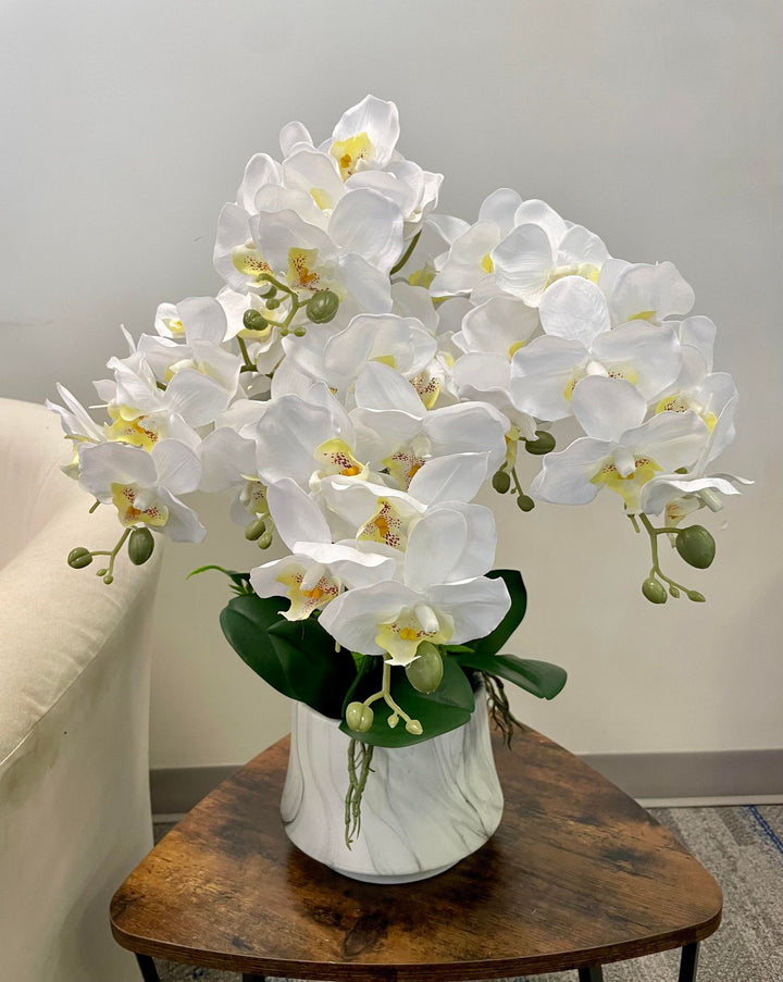 Arrangement of white orchids with delicate petals and yellow centers placed in a sleek white vase with marble-like patterns. The vase is set on a rustic wooden table, adding an elegant touch to the room's décor.