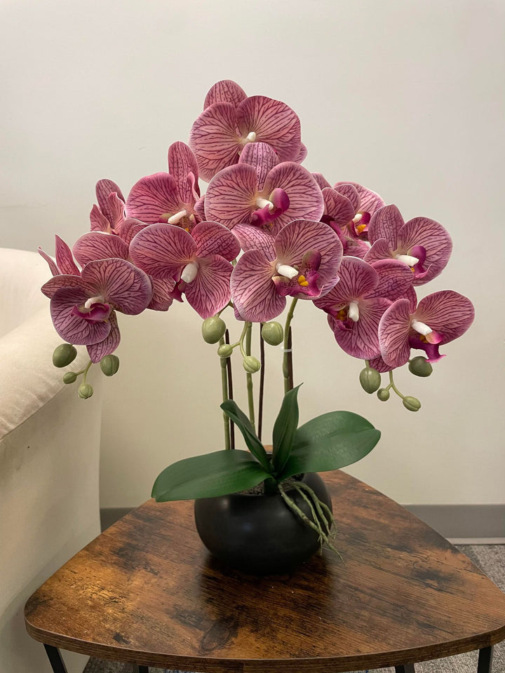 A beautiful arrangement of pink artificial orchids in a black planter, placed on a wooden side table. Frequently asked question: What are the best care tips for maintaining artificial orchids?