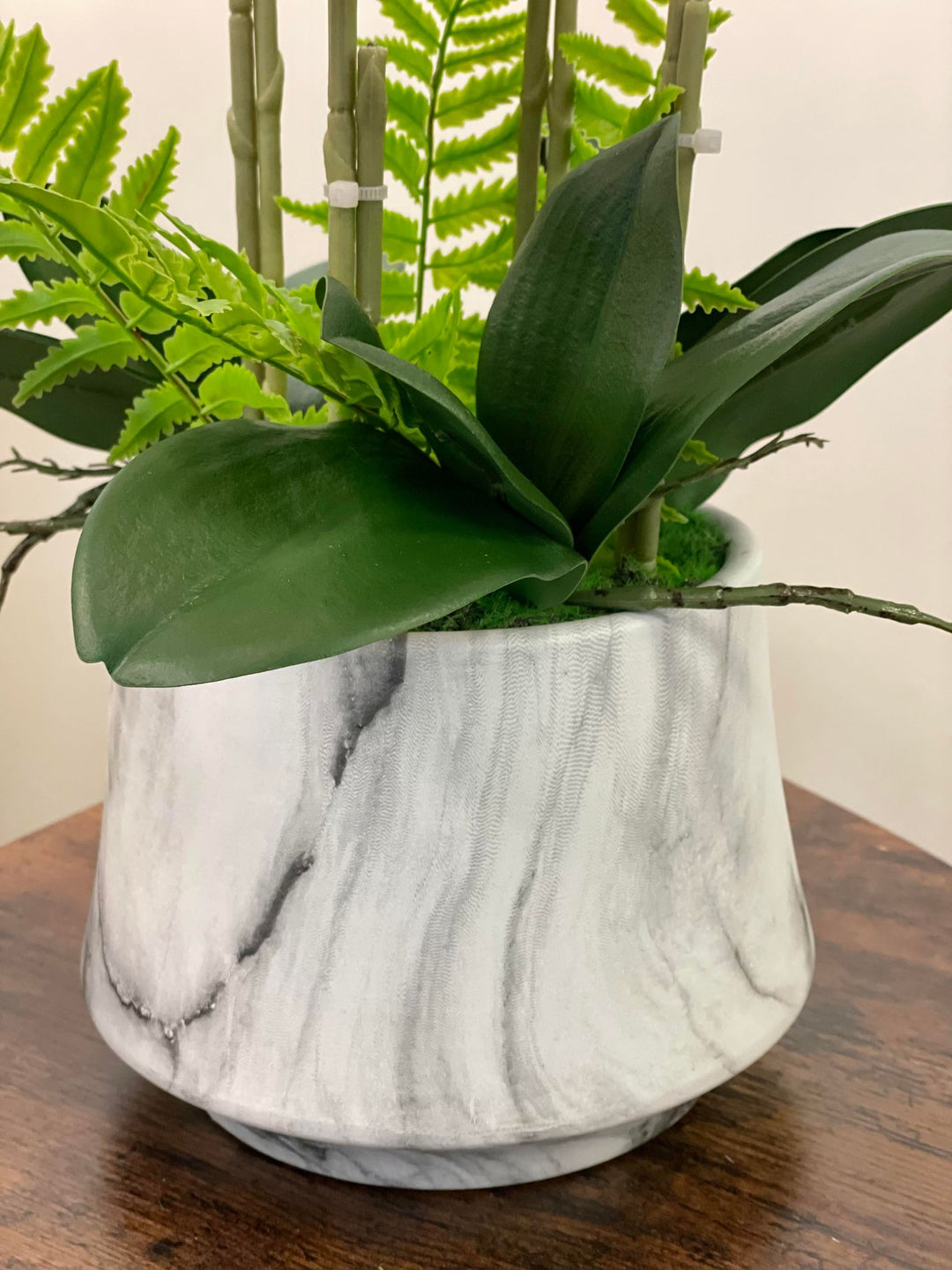 A close-up of a decorative plant arrangement featuring vibrant green leaves and bamboo stems placed in a marble-patterned vase. The vase is set on a wooden surface, highlighting the contrast between the sleek, modern design and the natural elements of the plant.