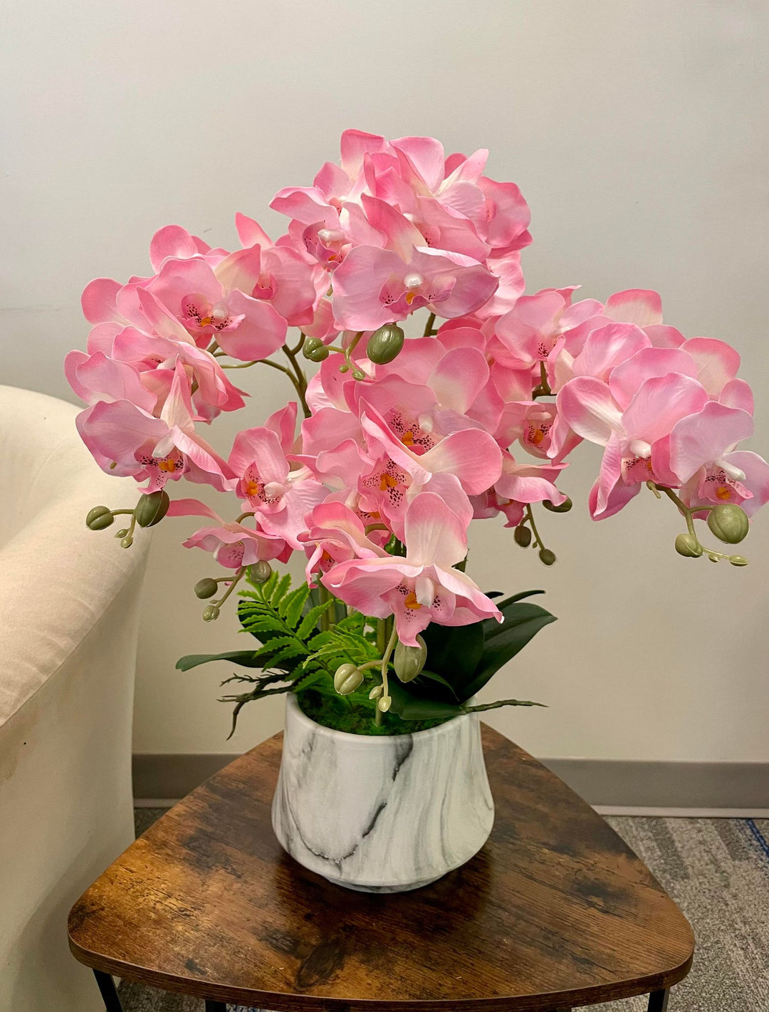 Pink orchids with delicate petals and yellow centers placed in a sleek white vase with marble-like patterns. The vase is set on a rustic wooden table, adding an elegant touch to the room's décor.