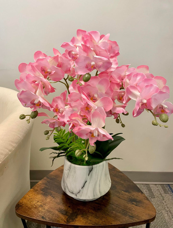 Pink orchids with delicate petals and yellow centers placed in a sleek white vase with marble-like patterns. The vase is set on a rustic wooden table, adding an elegant touch to the room's décor.