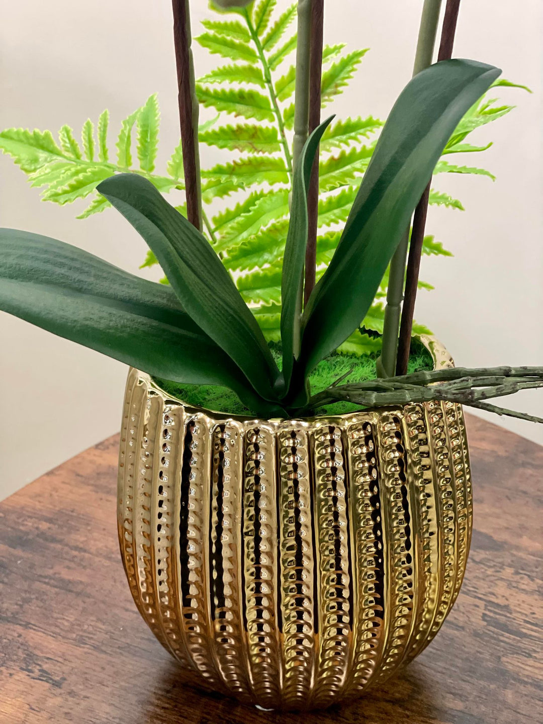 A close-up of a golden textured planter containing artificial orchid leaves and green ferns. Frequently asked question: What are the best pots or planters for displaying artificial plants indoors?"