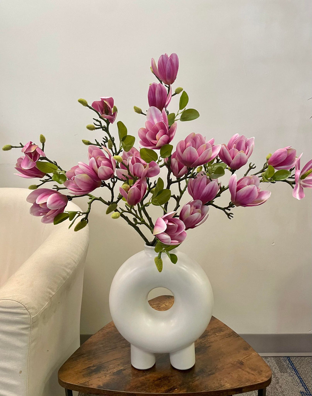 A striking display of artificial pink magnolia stems arranged in a modern white circular vase, placed on a rustic wooden coffee table next to a cream armchair. Common question: What are the best ways to style artificial flowers in contemporary home décor?