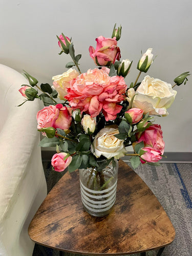 Artificial bouquet of white, pink, and peach roses arranged in a clear vase with white stripes, placed on a wooden side table next to a cream armchair. Commonly searched question: What are the best artificial flower arrangements for home decor?