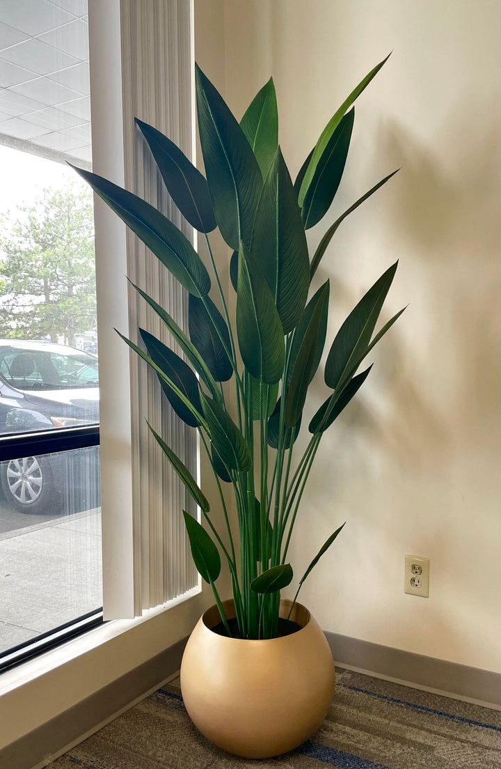 A tall artificial bird of paradise plant in a gold round planter positioned by a large window. The plant features realistic, elongated green leaves, adding a touch of natural elegance to the modern indoor space. The room's neutral tones highlight the vibrant green of the plant, making it an eye-catching decor element
