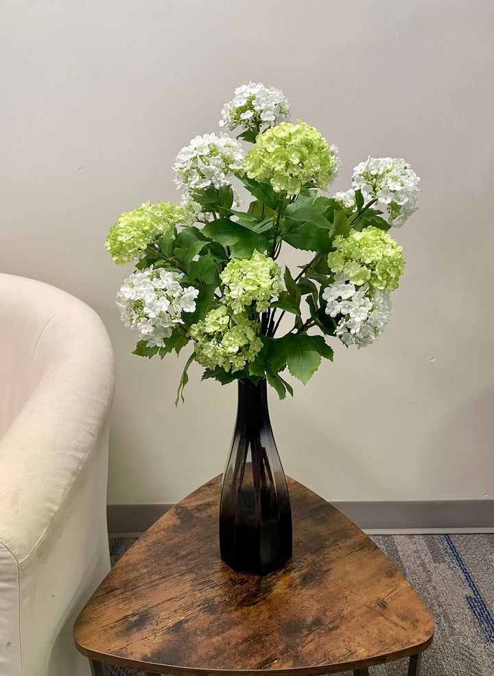 A black glass vase with a bouquet of green and white hydrangea artificial flowers placed on a wooden coffee table. Frequently searched question: How do you style artificial flowers in a vase for home decor?