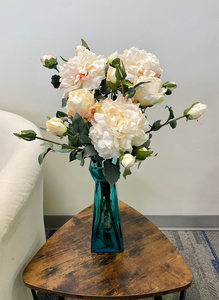 A bouquet of artificial white peonies arranged in a teal vase, sitting on a small wooden coffee table next to a cream-colored chair. Frequently asked question: How do I style artificial peonies in vases for a modern home décor setting?