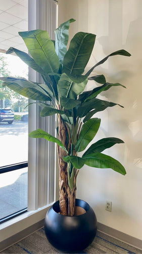 Artificial banana plant with large, vibrant green leaves, displayed in a modern black planter near a window in a well-lit indoor space