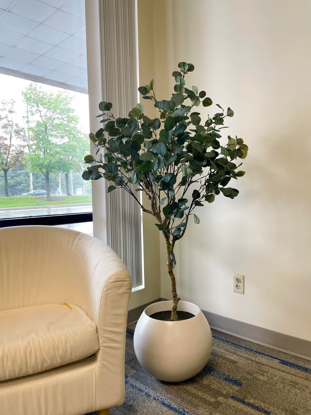 Artificial plant with broad, green leaves displayed in a modern, round white pot, placed next to a beige armchair, adding a natural touch to the indoor setting.