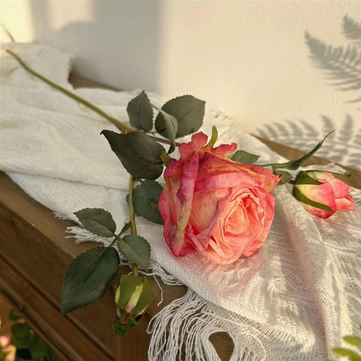 Artificial pink rose with green leaves lying on a white fabric, placed on a wooden surface. Frequently asked question: What are the best ways to decorate with artificial flowers for a realistic touch?