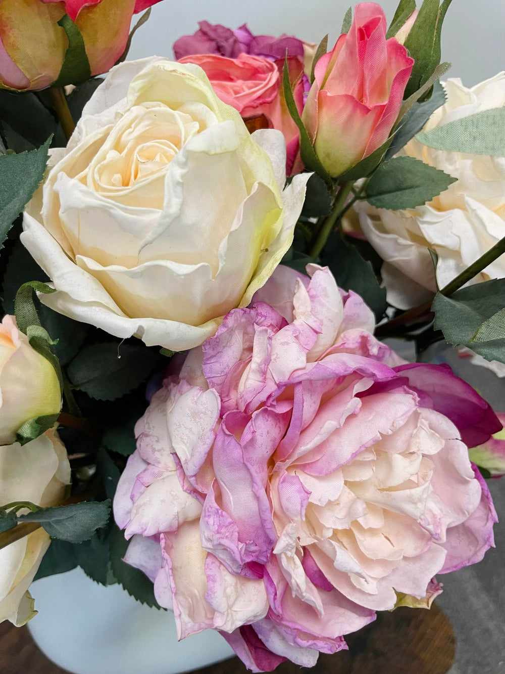 A close-up of artificial roses in soft shades of cream, pink, and lavender. The flowers showcase realistic textures with lush green leaves, arranged in a natural, lifelike bouquet.