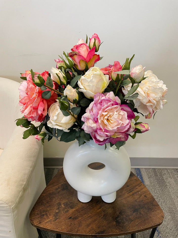 A beautiful arrangement of artificial roses in soft shades of purple, peach, and cream displayed in a white vase. The flowers are placed on a rustic wooden table next to a white armchair, creating a cozy, inviting corner.