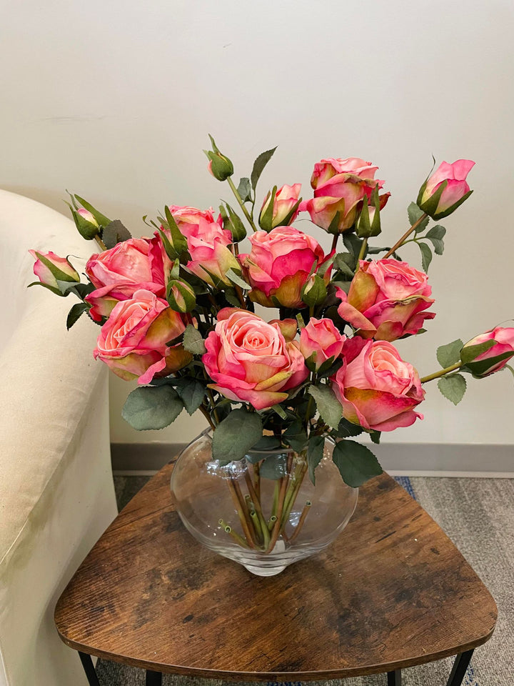 A beautiful arrangement of artificial pink and peach roses in a clear glass vase, placed on a rustic wooden table. The roses are surrounded by green leaves, adding elegance and charm to any room's décor.