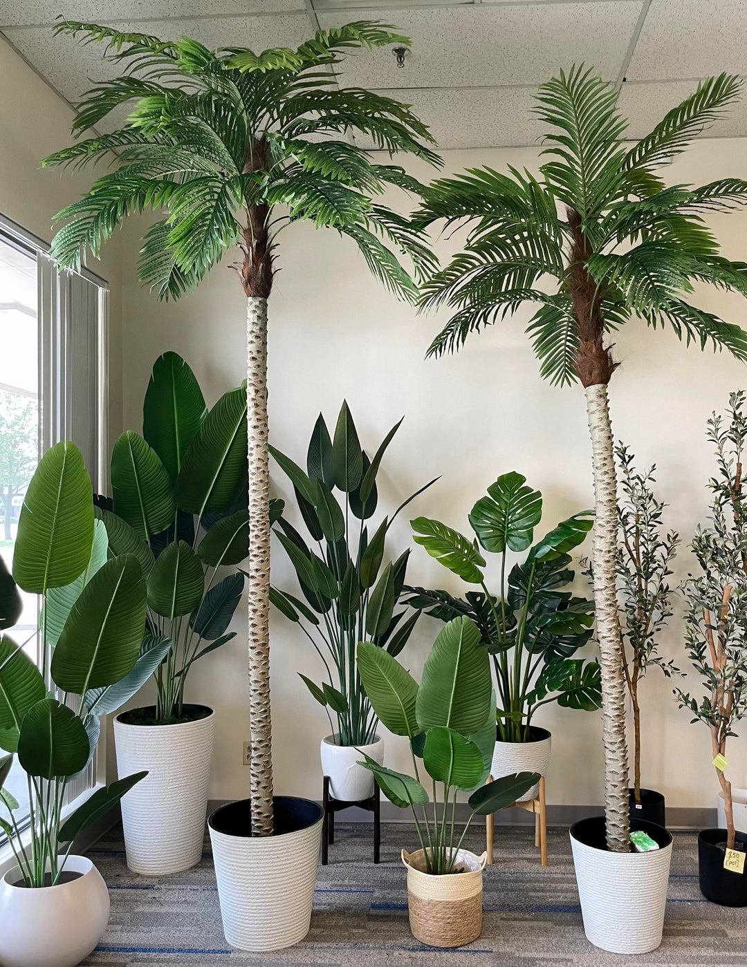 Indoor display of various artificial tropical plants, including two tall palm trees and a variety of potted plants, perfect for creating a lush, green ambiance in office or home interiors.