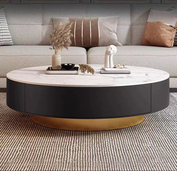 Elegant round coffee table with a white marble top and a black base featuring a gold accent at the bottom. The table is styled with decorative items and placed in front of a beige sofa with patterned pillows, creating a luxurious and modern living room setting.