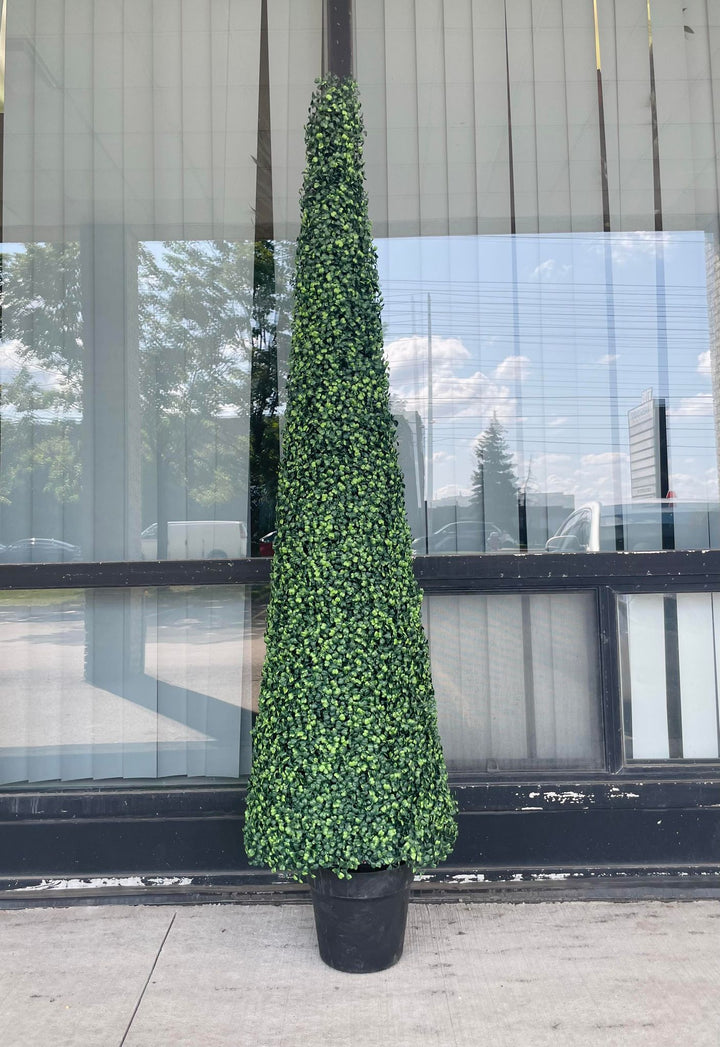A tall, artificial topiary in the shape of a cone stands in a black pot on a gray carpet. The topiary is positioned next to a white upholstered chair, providing a contrast between the soft texture of the chair and the dense, leafy surface of the topiary