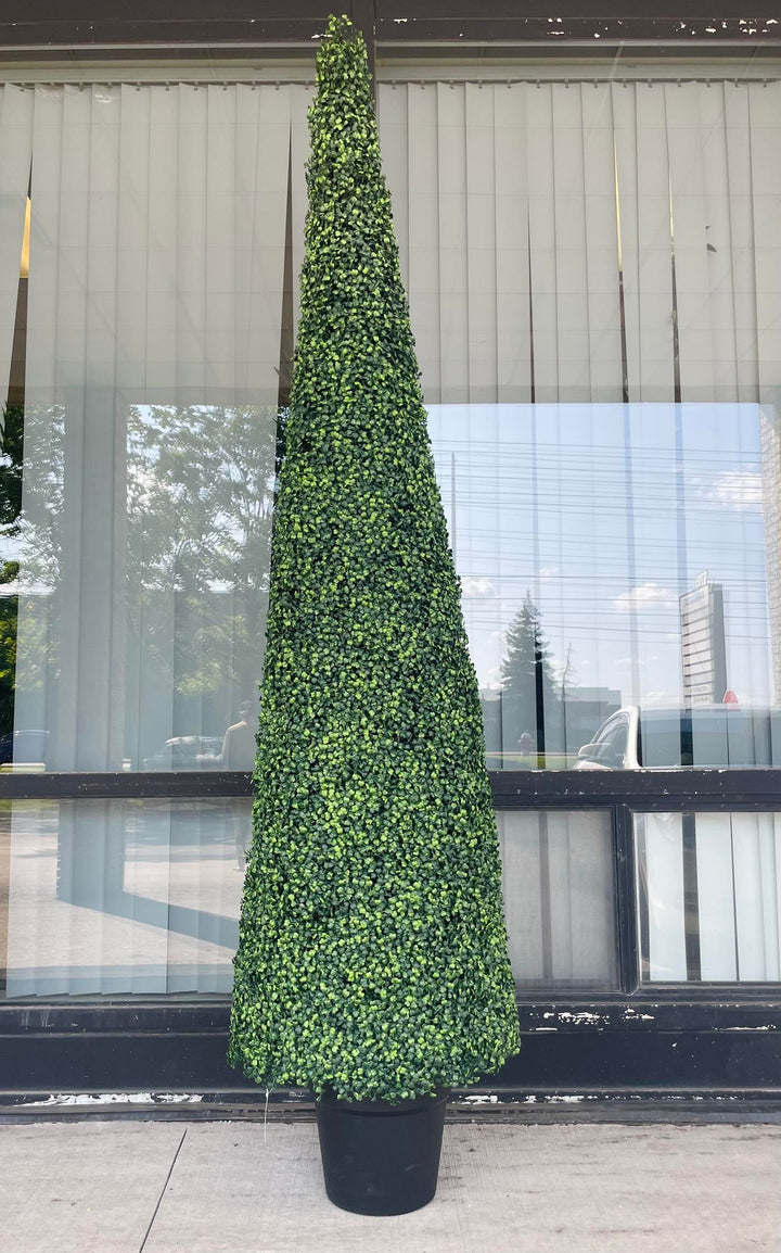 A tall, artificial topiary in the shape of a cone stands in a black pot. The topiary is lush with dense, small green leaves and is placed outside against a textured concrete wall. In the background, there is a view of a building with large windows