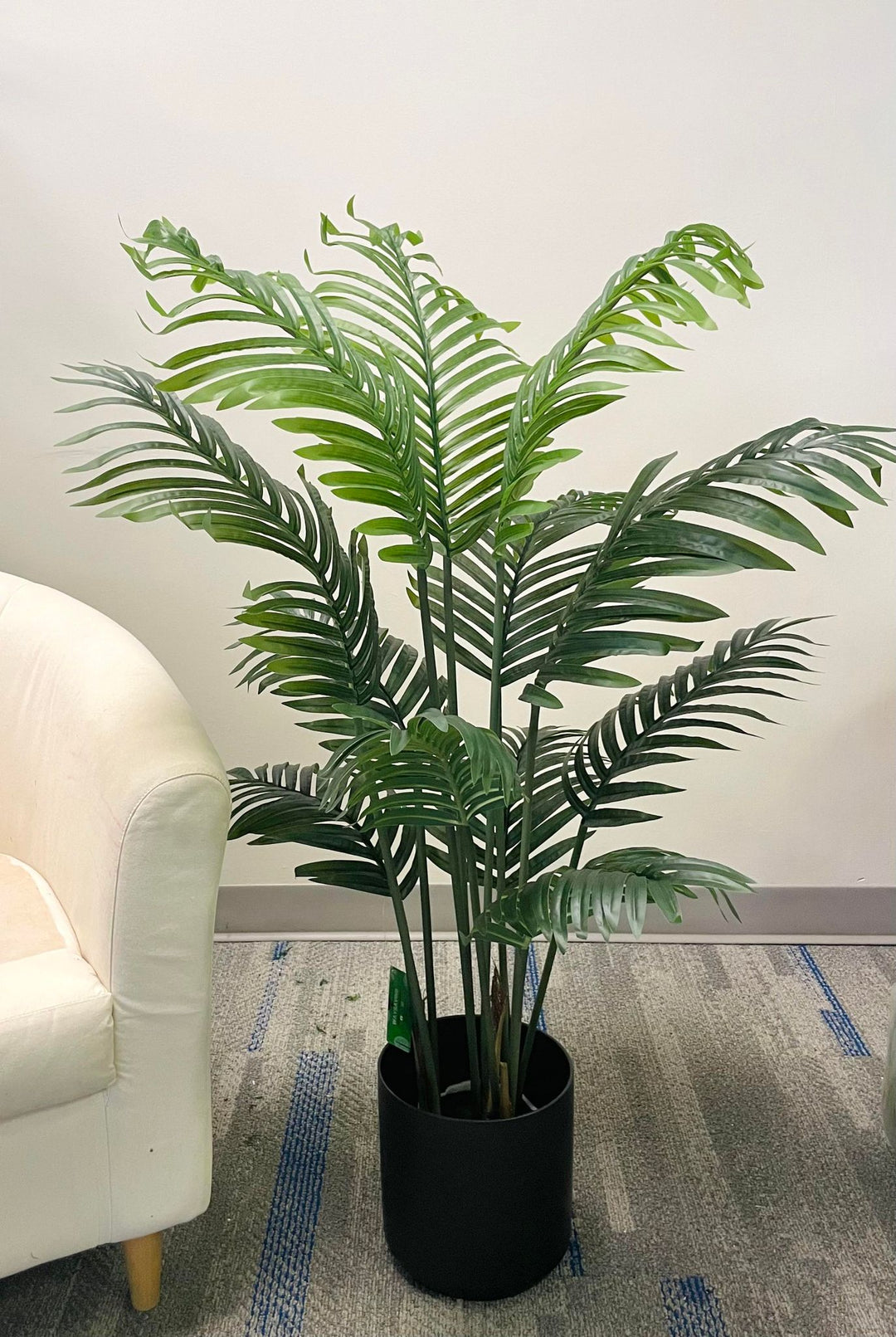 Artificial palm tree in a black planter, placed next to a cream armchair, creating a fresh and modern indoor décor setting.