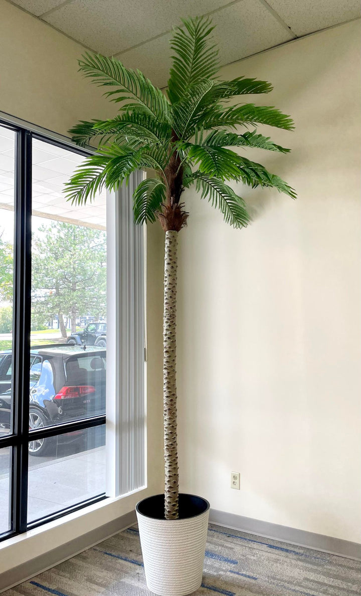 Artificial palm tree in a large, white textured planter, placed in a bright office space by the window, bringing tropical vibes and greenery to the indoor setting.