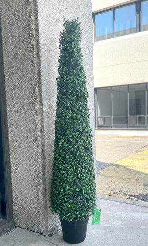 A tall, artificial topiary in the shape of a cone stands in a black pot. The topiary is lush with dense, small green leaves and is placed outside against a textured concrete wall. In the background, there is a view of a building with large windows.