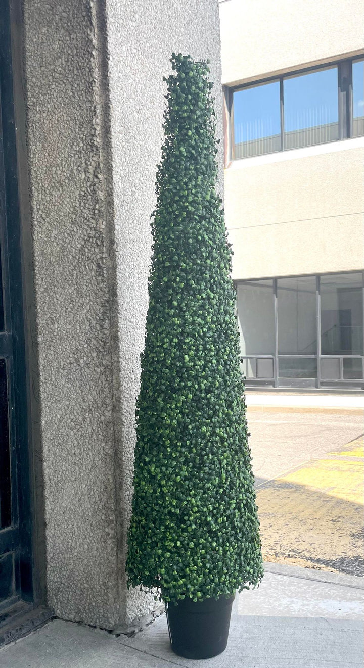 A tall, artificial topiary in the shape of a cone stands in a black pot. The topiary is lush with dense, small green leaves and is placed outside against a textured concrete wall. In the background, there is a view of a building with large windows.