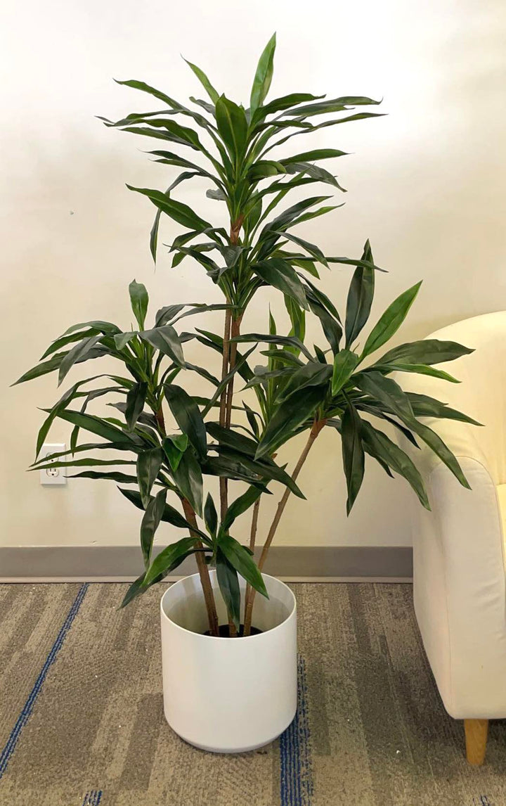 Artificial green Dracaena plant in a modern white planter with wooden stand, placed in a well-lit room next to a sofa, adding a natural and elegant touch to the indoor space.