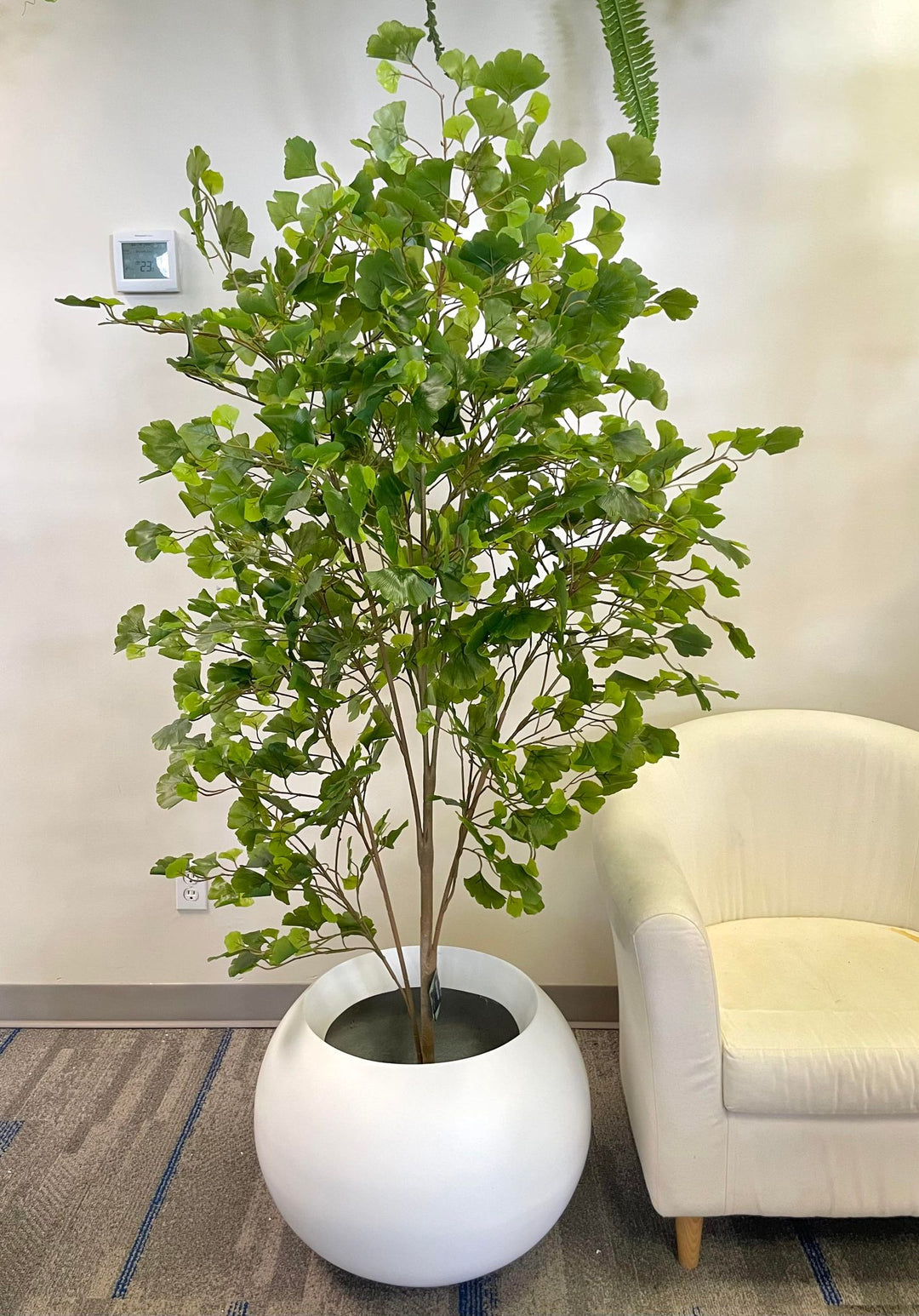 Artificial tree in a round white planter placed next to a cream-colored chair, enhancing the indoor space with natural-looking greenery.