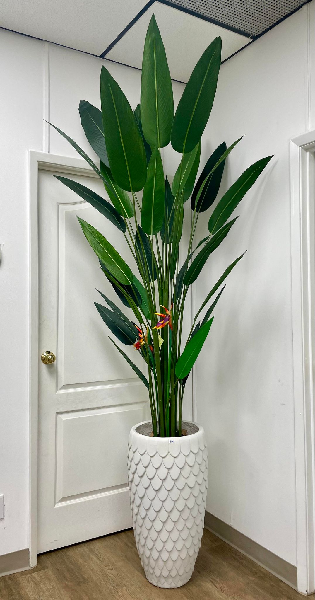 Tall artificial bird of paradise plant with large green leaves and vibrant orange flowers, set in a textured white ceramic planter. The plant is positioned indoors against a white wall and door, adding a touch of greenery to the space.
