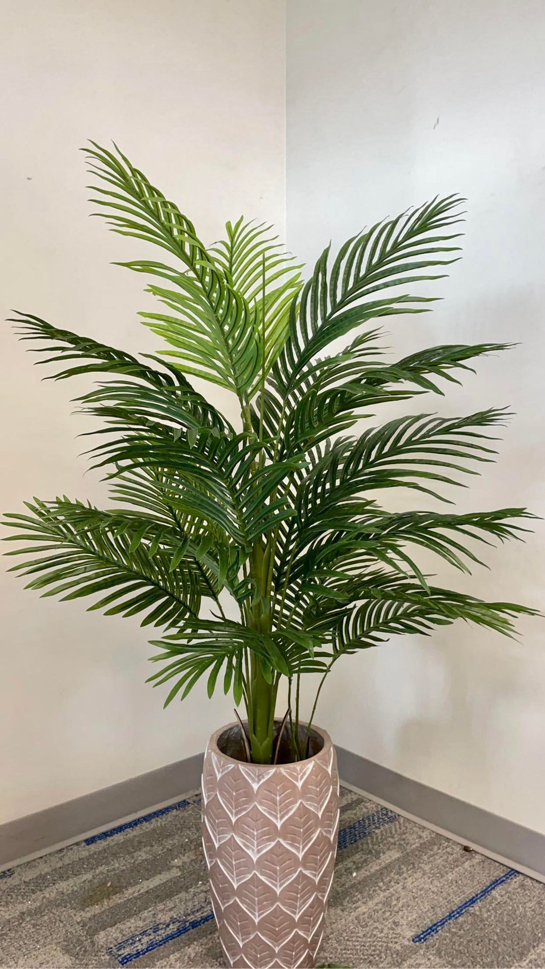Artificial palm tree in a decorative pot with detailed green leaves displayed indoors against a beige wall.