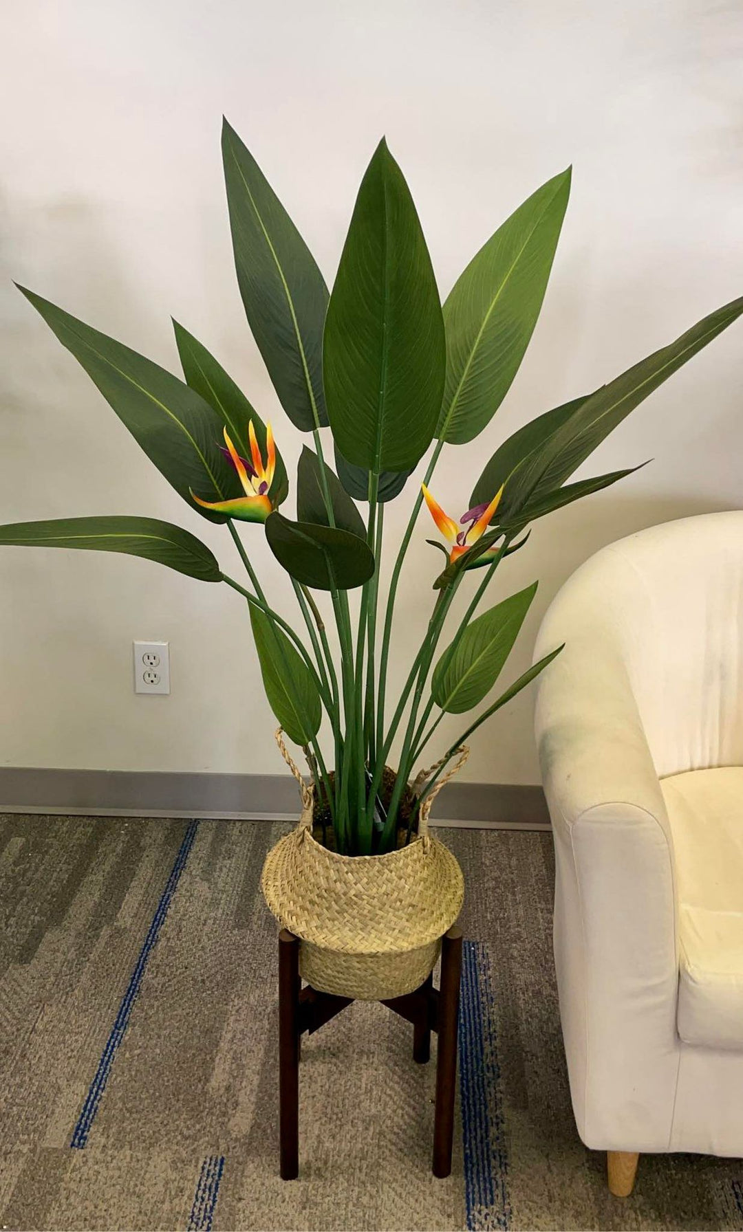 Artificial Bird of Paradise plant with large green leaves and bright orange and purple flowers, placed in a woven basket on a wooden stand next to a white chair.