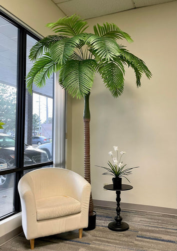 Modern office corner featuring a tall artificial palm tree, a cozy white armchair, and a black side table with a minimalist white floral arrangement, creating a welcoming and serene environment.